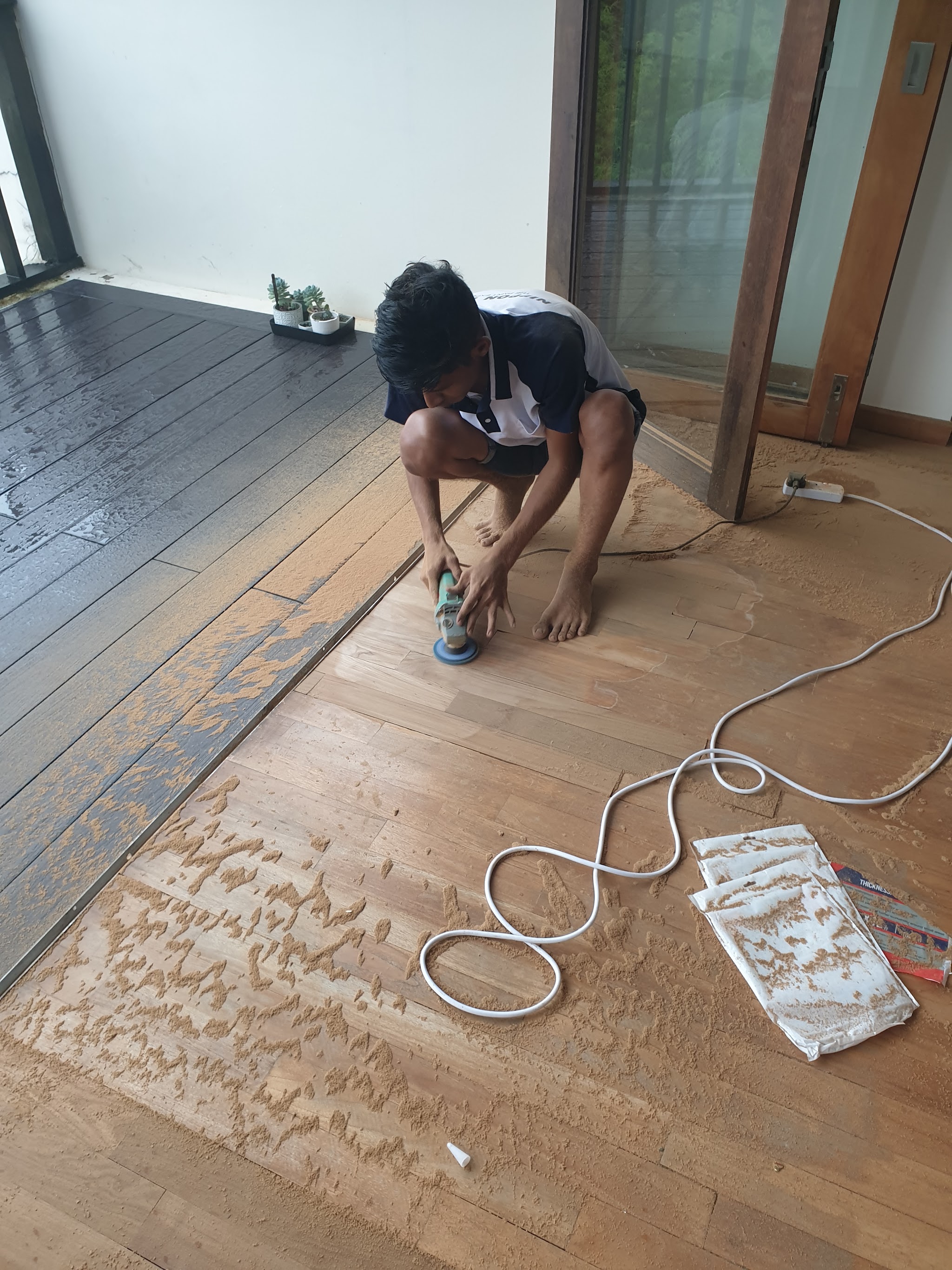 A tradesman kneeling to sand a timber floor with a small rotary sander, sawdust across the boards